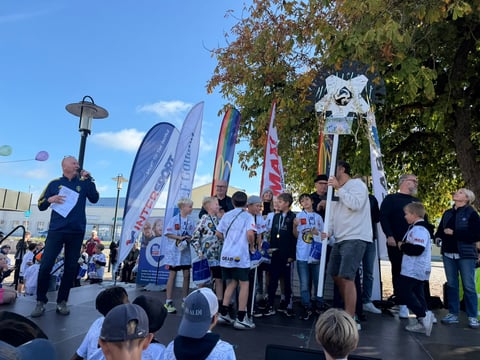 Outdoor community event with crowds gathered around a stage, speaker at podium, promotional banners, and clear blue sky with trees in background