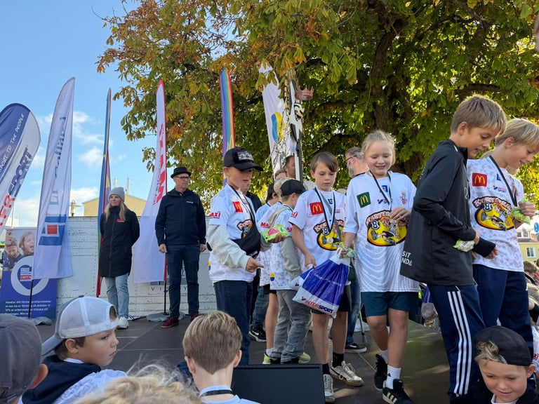 Children wearing medals stand on a podium at an outdoor sports event with banners and trees in the background