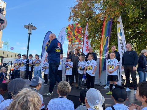 Outdoor community event with uniformed group on stage, colorful decorations, and crowd viewing under blue sky