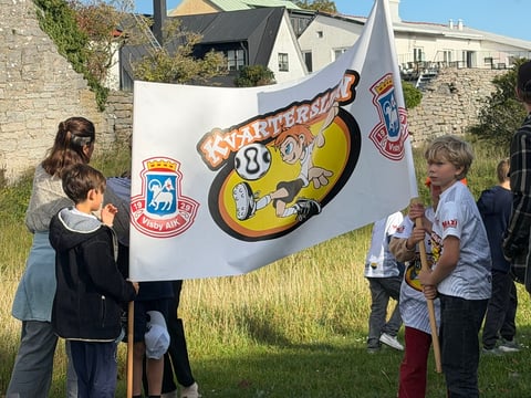 Children holding a vintage skateboard-themed banner outdoors in a grassy area with stone buildings in background