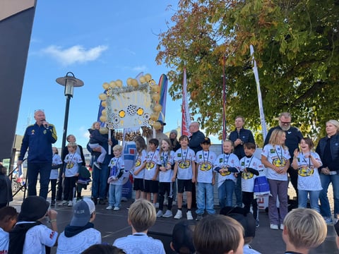 Youth baseball team and coaches on stage during outdoor victory celebration with crowd watching