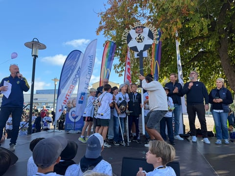 Outdoor community event with people gathered on a stage beneath banners and flags on a sunny day