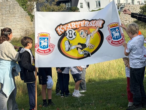 Group of children holding a large banner with cartoon mascot and logos at an outdoor event