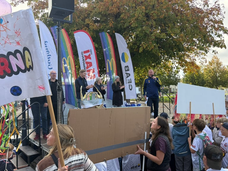 Outdoor event with large branded banners and crowds gathered around a stage or presentation area