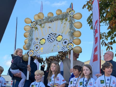 Group of children standing beneath large decorative frame with golden spheres and ornaments hanging from it at outdoor event