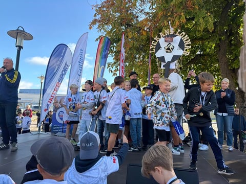 Group of participants in blue and white shirts standing on an outdoor stage with banners and trees, outdoor event or fundraiser