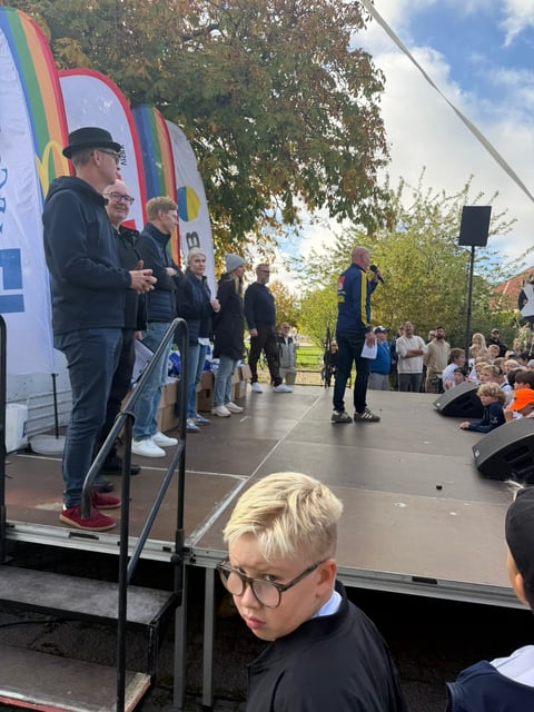 Outdoor community event with speakers on stage featuring rainbow Pride flag decorations and seated audience