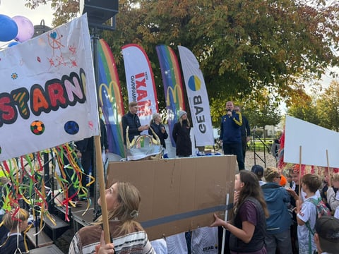 Children participate in an outdoor carnival or festival with colorful carnival rides and game booths surrounded by autumn trees