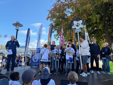 Group of children and adults on outdoor stage at community event with blue flags and banners, clear blue sky and large tree in background