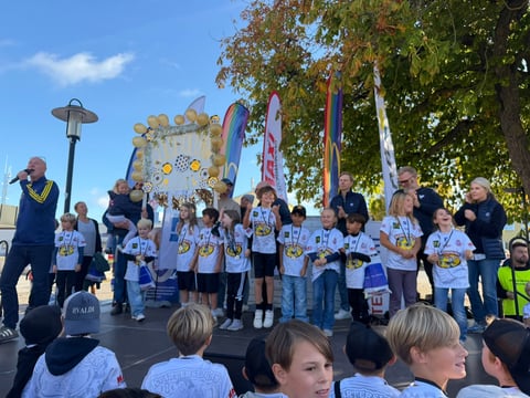 Group of children in blue and white team shirts standing on stage at an outdoor community event with banners and trees in background